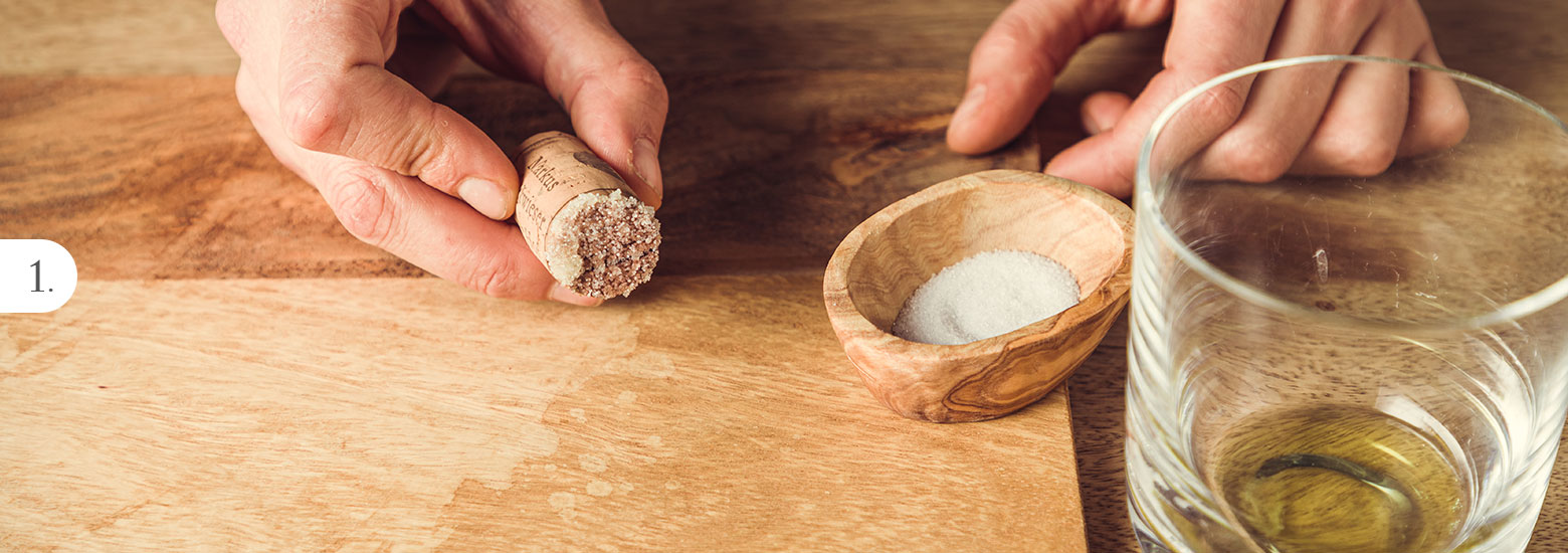 Eine Person zeigt einen Korken, der in Möbel-Öl und in Salz getaucht wurde. Im Vordergrund: Schälchen mit Salz und Glas mit Öl.