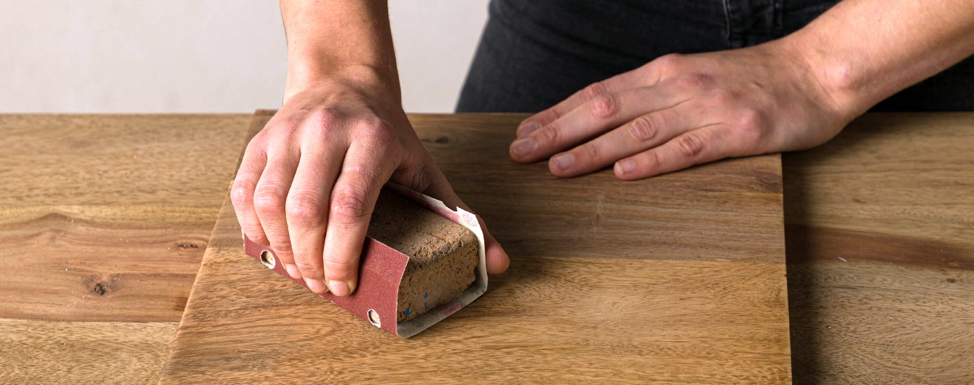 Eine Person schleift ein Holzbrett mit einem Schleifblock per Hand ab.